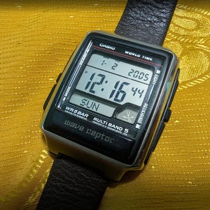 May include: A Casio Wave Ceptor digital watch with a black leather band. The watch face displays the time as 12:16:44, the date, and the day of the week. The watch has a silver and black case.