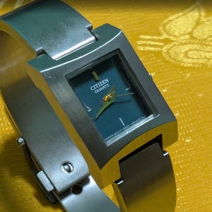 Könnte beinhalten: Eine Citizen Quartz Armbanduhr aus Edelstahl mit quadratischem Gehäuse und dunkelblaugrünem Zifferblatt. Die Uhr hat goldfarbene Zeiger und ein breites, integriertes Metallarmband mit Verschluss. Auf dem Zifferblatt steht Citizen Quartz.
