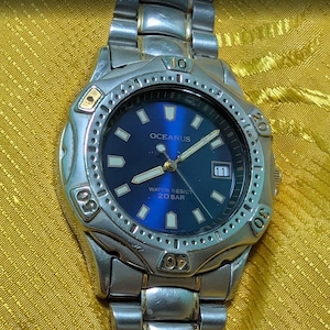 May include: A silver-tone stainless steel analog wristwatch with a deep blue circular dial, luminous hour markers, and hands. The watch features a rotating bezel with engraved numbers, a date window at the three o'clock position, and the text OCEANUS.