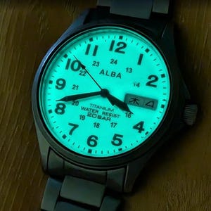 May include: An Alba wristwatch with a dark metal case and bracelet, featuring a luminous light-green dial that glows in the dark. The face displays black Arabic numerals, a 24-hour scale, a date window, and the text Titanium Water Resist 20 Bar.