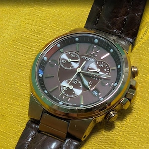 May include: A Citizen chronograph watch with a brown dial, three sub-dials, and a date window. The watch features a gold-tone metal case and a dark brown textured leather strap with visible stitching. The dial includes luminous hands and markers.