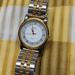 May include: A Seiko Lucent wristwatch featuring a round white dial with Roman numeral hour markers, a gold-tone bezel, and a two-tone silver and gold metal link bracelet. The watch face displays the Seiko brand name and the Lucent model name.