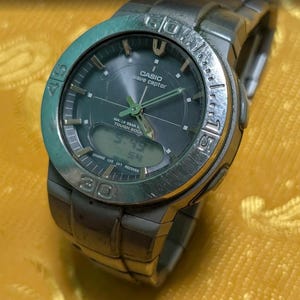 May include: A silver Casio Wave Ceptor watch with a dark green face and digital display. The watch has a silver band and a rotating bezel with numbers. The time displayed is 3:49.