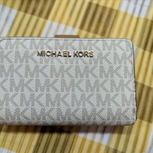 Op de afbeelding: Een kleine, rechthoekige Michael Kors portemonnee. De portemonnee is wit met een herhalend patroon van de letters "MK" in lichtbeige. De woorden "MICHAEL KORS" zijn in gouden letters op de voorkant gedrukt.