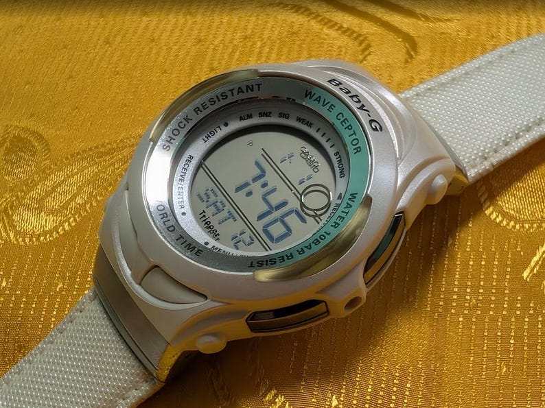 May include: A white Baby-G Wave Ceptor digital watch with a light green bezel and a matching white strap. The watch face displays the time, date, and other functions. The watch is shock resistant and water resistant.