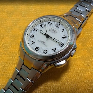 Pode incluir: Um relógio de pulso Citizen Eco-Drive Attesa em tom prateado com mostrador branco texturizado, algarismos arábicos pretos e ponteiros luminosos. O relógio possui uma pulseira de elos de aço inoxidável, caixa redonda e tecnologia de rádio controle.