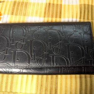 May include: Black leather wallet with embossed repeating "Dior" logo pattern. The wallet is rectangular and has a fold-over flap. The background is a yellow and white striped fabric.