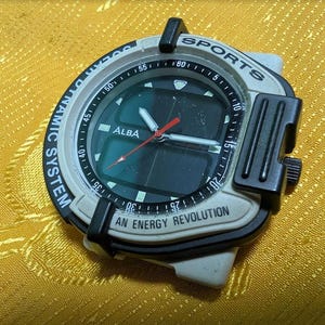 May include: A vintage Alba Sports watch with a black and white face, featuring a digital display and red second hand. The watch has a black bezel and a gray case, with the text "AN ENERGY REVOLUTION".
