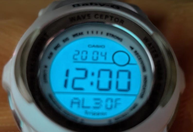 May include: A white Casio Baby-G Wave Ceptor digital watch with a blue illuminated display. The display shows the time as 12:00, date as 20/04, and other indicators. The watch has a round face and a white band.