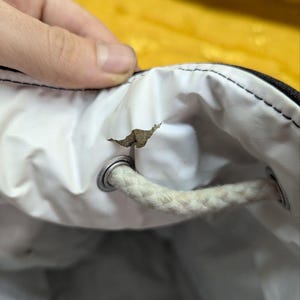 May include: Close-up of a white drawstring bag interior with a torn section. The bag has a black exterior and a white interior. A white rope drawstring is visible, along with a metal grommet.