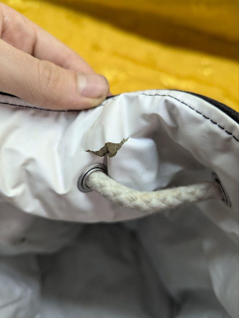 May include: Close-up of a white drawstring bag interior with a torn section. The bag has a black exterior and a white interior. A white rope drawstring is visible, along with a metal grommet.