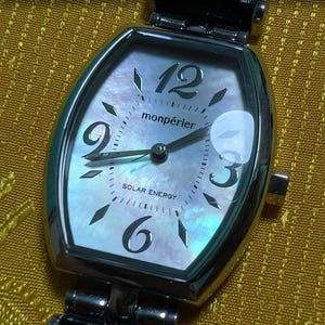 Può includere: Un orologio da polso argentato con quadrante in madreperla. L'orologio ha una cassa rettangolare con bordi arrotondati e un cinturino nero. Il quadrante mostra il marchio "monpèrier", le parole "solar energy" e i numeri 12, 2, 3 e 6.