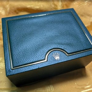 Pode incluir: Uma caixa de relógio azul escuro com um acabamento texturizado semelhante ao couro. A caixa tem uma moldura prateada e um logotipo de coroa prateada na frente. A caixa é retangular e está sobre uma superfície dourada.