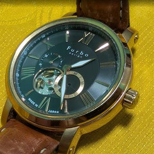 May include: A Furbo design wristwatch with a gold-tone metal case, a dark dial featuring Roman numerals, and a brown leather strap. The watch face displays an open-heart movement, a sub-dial, and the text Furbo design and Made in Japan.
