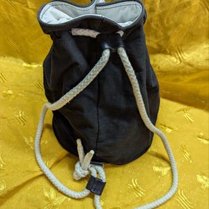 May include: A black drawstring bag with a white rope closure. The bag has a white interior and a black leather-like accent. The rope is white and knotted at the bottom. The bag is made of a dark fabric.