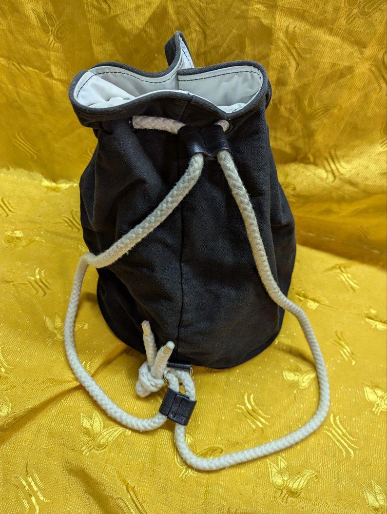 May include: A black drawstring bag with a white rope closure. The bag has a white interior and a black leather-like accent. The rope is white and knotted at the bottom. The bag is made of a dark fabric.