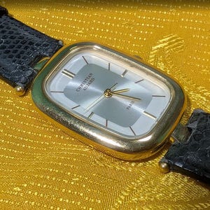 Op de afbeelding: Een goudkleurig horloge met een rechthoekige wijzerplaat en een zilveren wijzerplaat. Het horloge heeft de tekst "CHRISTIAN AUJARD PARIS" en heeft gouden wijzers en markeringen. Het horloge heeft een zwarte leren band met een textuur.