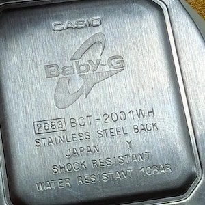 May include: Close-up of the back of a Casio Baby-G watch. The watch back is stainless steel and features the Baby-G logo, model number 2683 BGT-2001WH, and text indicating it is shock and water resistant.