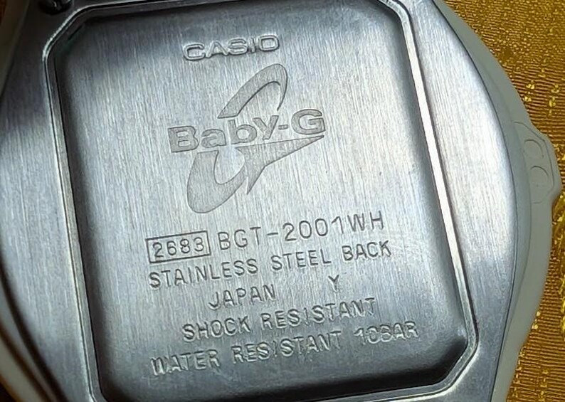 May include: Close-up of the back of a Casio Baby-G watch. The watch back is stainless steel and features the Baby-G logo, model number 2683 BGT-2001WH, and text indicating it is shock and water resistant.