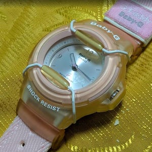 Puede incluir: Un reloj Baby-G Casio con una correa rosa claro y una caja naranja transparente. La esfera del reloj es plateada con números y manecillas negras. El reloj está etiquetado como "Shock Resist" y "Water Resist".