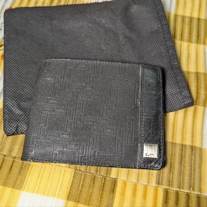 Puede incluir: Una cartera de cuero negro con un patrón geométrico y un pequeño logotipo plateado. La cartera está abierta, revelando el interior. Una bolsa gris oscuro está detrás de la cartera. Los artículos están sobre una superficie a rayas amarillas y blancas.