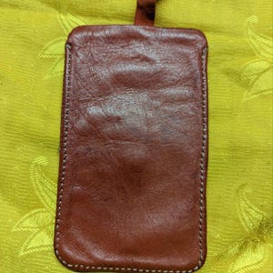 May include: A rectangular, reddish-brown leather case with a stitched border and a small, attached red strap. The case is likely designed to hold a phone or other small items. The background is a textured, yellow-green fabric.