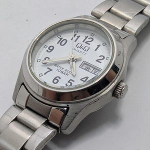 Pode incluir: Um relógio de pulso em aço inoxidável prateado com mostrador branco, algarismos arábicos pretos e ponteiros prateados. O mostrador apresenta o texto Q&Q QUARTZ, WATER RESIST 10 BAR e uma janela de dia e data. Inclui pulseira de elos metálicos.