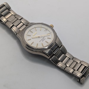 Op de afbeelding: Een Seiko solar titanium polshorloge met een zilverkleurige metalen schakelband en een ronde witte wijzerplaat. Het horloge heeft goudkleurige wijzers en uurmarkeringen, een datumvenster op de drie-uurpositie en een kleine gouden kroon.