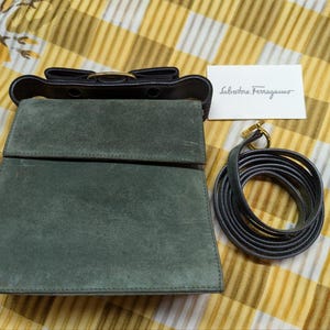 May include: A green suede handbag with a black leather top and a gold-tone clasp. A matching black leather strap is coiled beside the bag. A small white card with the text "Salvatore Ferragamo" is also present.