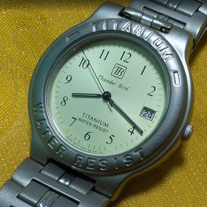 Könnte beinhalten: Eine silberfarbene Titanium Thunder Bird-Uhr mit cremefarbenem Zifferblatt. Die Uhr hat schwarze Zeiger, eine Datumsanzeige und die Worte "TITANIUM" und "WATER RESIST" auf der Lünette. Das Armband ist ebenfalls silberfarben.