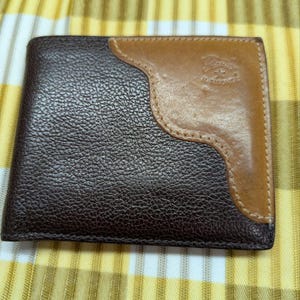 May include: A bi-fold leather wallet in dark brown with a tan accent. The wallet has a textured surface and a stitched border. The tan accent features a logo. The wallet is closed and rectangular.