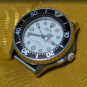 May include: A silver-tone wristwatch with a white dial, circular hour markers, and a black rotating bezel featuring white numerals. The dial displays the text OXY, VEGA, and WATER 200M RESIST, with a day-date window positioned at the three o'clock mark.