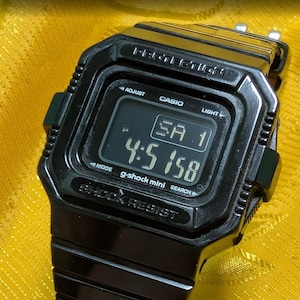 Puede incluir: Un reloj digital negro G-Shock Mini con una esfera rectangular. El reloj muestra la hora 4:51:58 y el día SA 1. El reloj tiene las palabras "SHOCK RESIST" y "PROTECTION" impresas.
