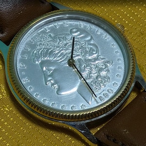 May include: A silver-toned watch with a coin face featuring a profile and the year 1890. The watch has a gold-colored bezel, a brown leather strap, and black hands. The watch face has the words "E PLURIBUS UNUM".