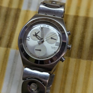 May include: A silver-toned Swatch chronograph watch with a metallic link bracelet. The watch face features a light silver dial, three sub-dials, and the Swatch Swiss logo. It includes three side buttons and Roman numeral markers at the twelve and nine positions.