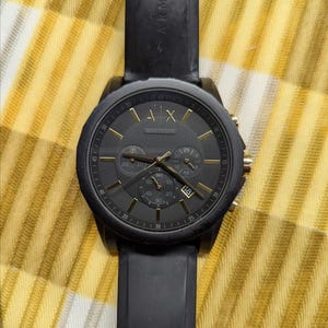 May include: Black Armani Exchange watch with a black band and a black face with gold-colored accents. The watch has three sub-dials and a date display. The watch is on a yellow and white checkered surface.