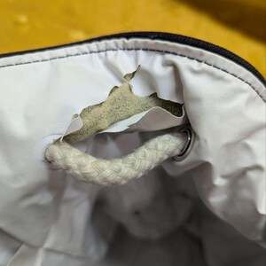 May include: Close-up of a white bag interior with a torn section revealing the inner padding. A thick, braided white rope handle is attached with a metal ring. The bag's exterior is black.