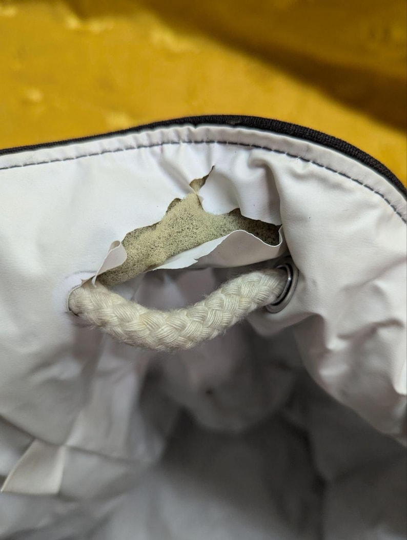 May include: Close-up of a white bag interior with a torn section revealing the inner padding. A thick, braided white rope handle is attached with a metal ring. The bag's exterior is black.