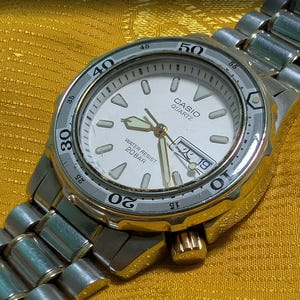 May include: A silver-toned Casio Quartz watch with a white face, featuring a date display and luminous hands. The watch has a rotating bezel with minute markers and a stainless steel band. The watch is water resistant to 20 bar.