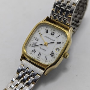 May include: A Claude Bernard quartz wristwatch with a gold-tone rectangular case and a white dial featuring black Roman numeral hour markers. The watch includes a date window at the three o'clock position and a two-tone silver and gold link bracelet.
