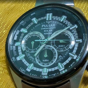 Könnte beinhalten: Eine silberfarbene Pulsar Solar-Chronograph-Armbanduhr mit schwarzem Zifferblatt, drei Hilfszifferblättern, leuchtenden Stundenmarkierungen und einer Tachymeterskala auf der Lünette. Das Zifferblatt zeigt den Text Pulsar, Chronograph, Solar und 100M.