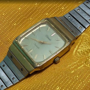 Puede incluir: Un reloj de cuarzo Clewaz vintage con una caja rectangular dorada y una correa de metal plateado. La esfera del reloj es de color claro con manecillas y marcadores negros. El reloj se muestra sobre una superficie dorada texturizada.