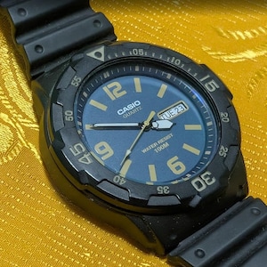 Pode incluir: Relógio Casio Quartz preto com mostrador azul, algarismos e ponteiros dourados. O relógio exibe o dia e a data, com o texto "WATER RESIST 100M". Possui uma moldura e pulseira pretas.