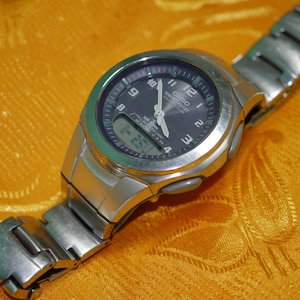 May include: A silver-tone Casio Wave Ceptor watch with a round metallic case and a matching link bracelet. The dark dial features white Arabic numerals, luminous hands, and a digital display window at the bottom showing the time as 1:25.