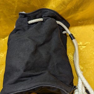 May include: Black drawstring bag with a white rope closure. The bag is made of a dark fabric and has a simple, casual design. The rope is thick and knotted at the end.