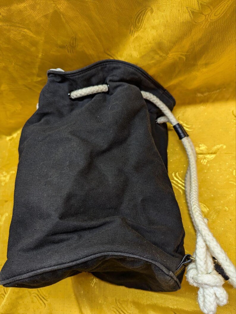 May include: Black drawstring bag with a white rope closure. The bag is made of a dark fabric and has a simple, casual design. The rope is thick and knotted at the end.