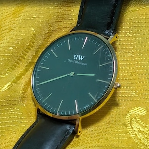 Puede incluir: Un reloj Daniel Wellington dorado con una correa de cuero negro. La esfera del reloj es verde oscuro con manecillas y marcadores dorados. El reloj está sobre un fondo amarillo texturizado.
