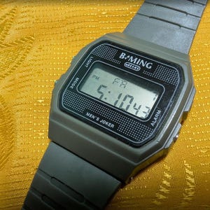 May include: Gray digital watch with a rectangular face displaying the time 5:10:43 PM. The watch has the text "B.MING" and "MEN'S JOKER" printed on the face. The watchband is gray and the background is yellow.