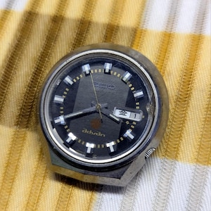May include: A round silver-tone Seiko Advan watch head with a dark gray dial, silver hands, and white hour markers. The dial features the text Seiko and Advan, along with a day-date window displaying the Japanese character for Friday and the number 1.