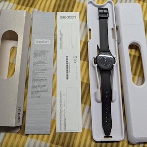 Puede incluir: Un reloj Swatch negro con una correa negra, presentado en su embalaje blanco original. La esfera del reloj es oscura y la correa se sujeta con una hebilla negra. El embalaje incluye una caja y documentos.